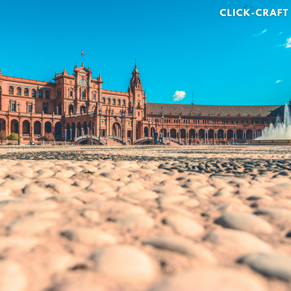 A grand, ornate building with intricate architecture and fountains in the foreground, set against a bright blue sky with clouds, Seville.
