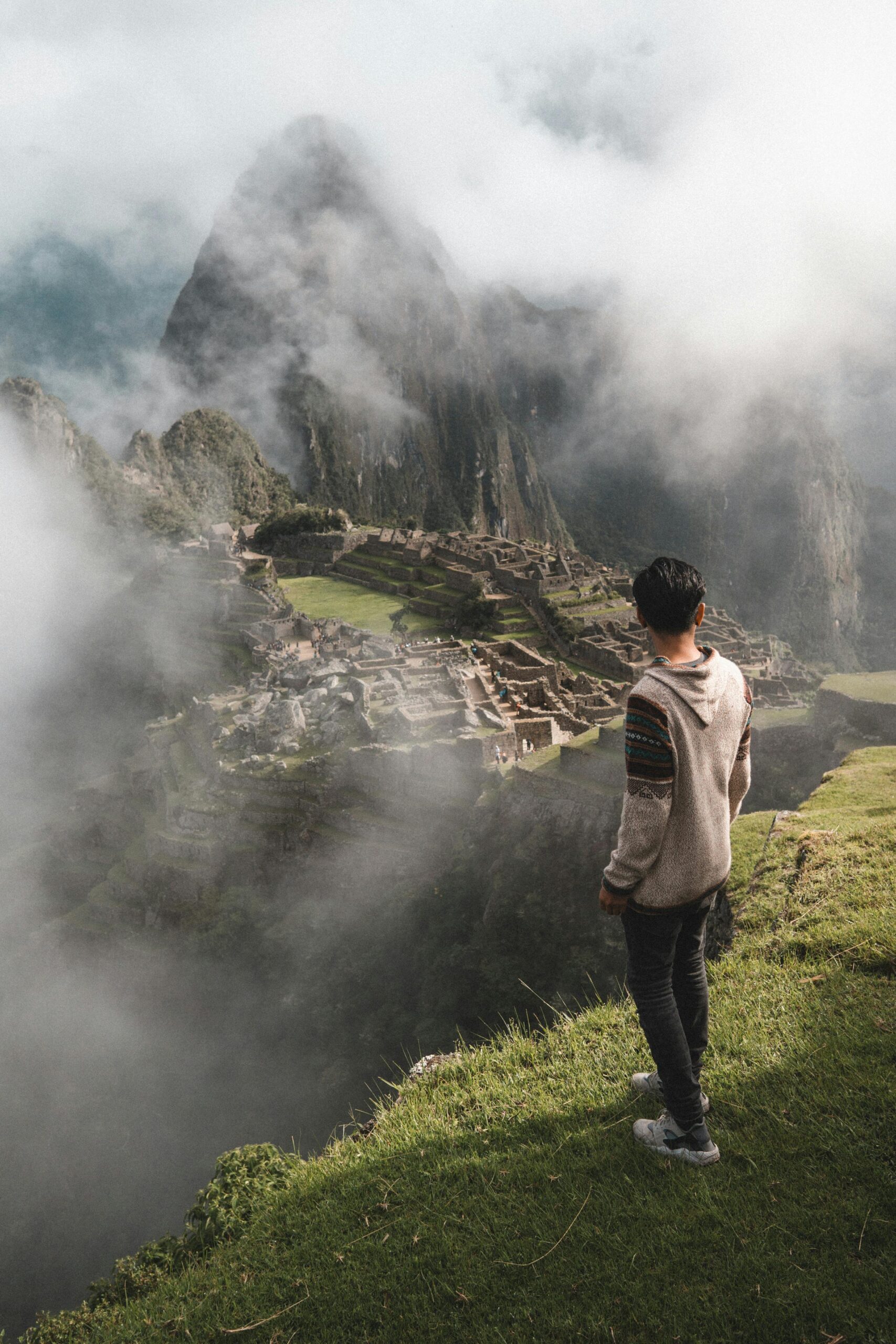 A person standing on a grassy hill, gazing out at the ancient ruins of Machu Picchu, partially obscured by mist and fog. Lima