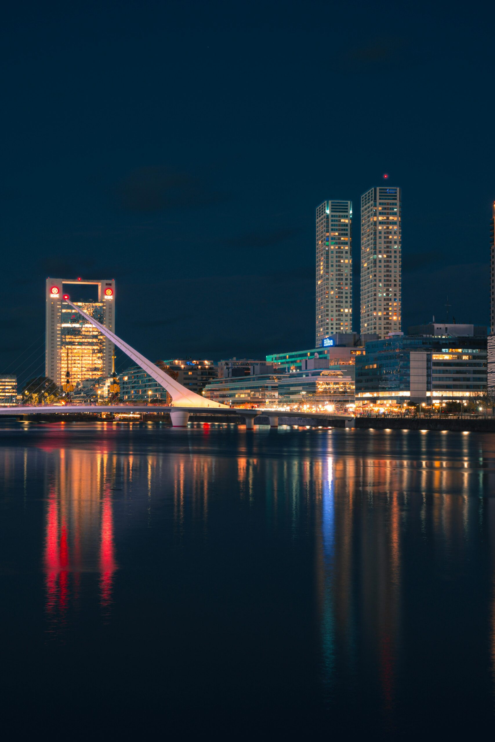 A nighttime cityscape with illuminated skyscrapers and a bridge reflected in the water. Buenos Aires