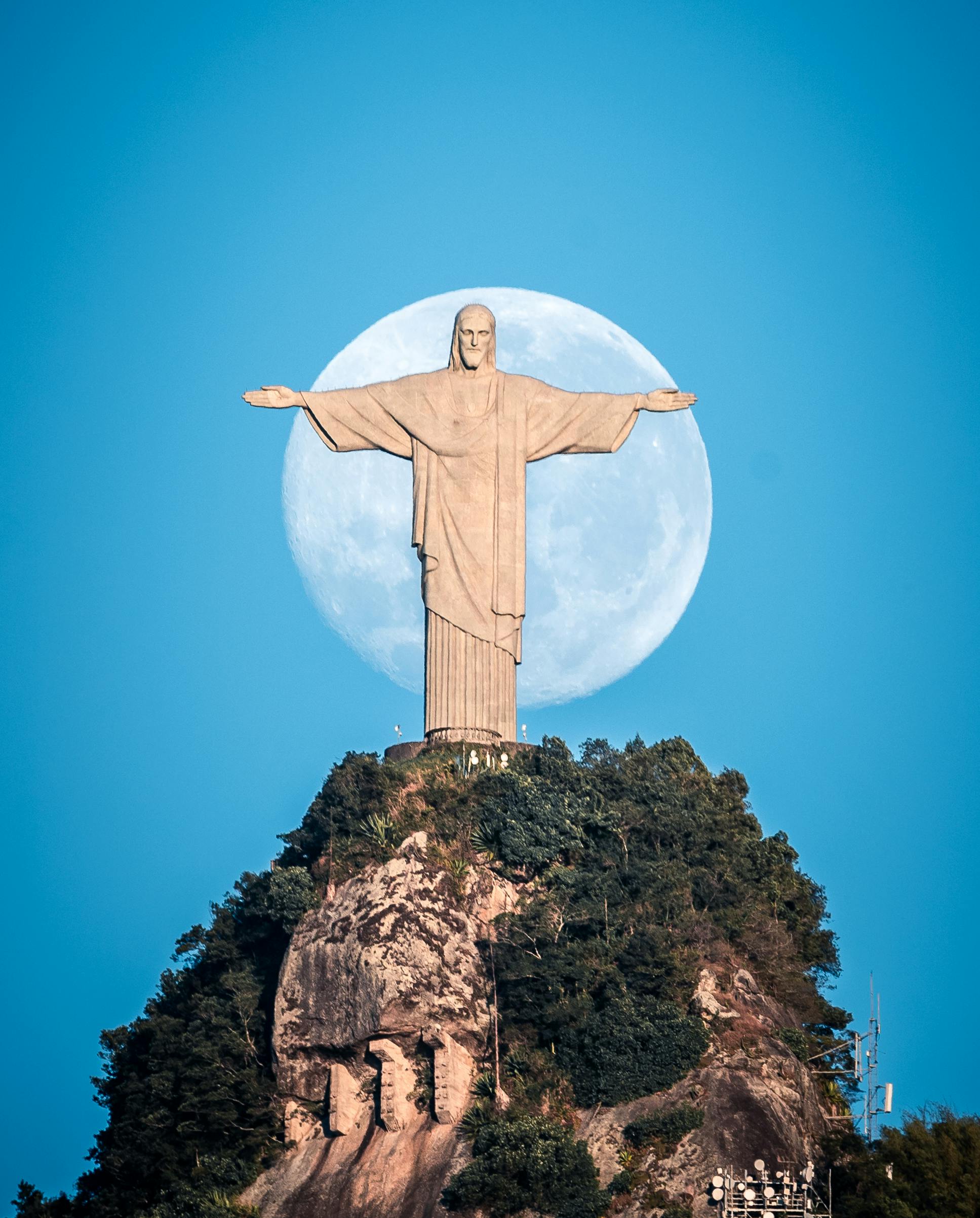 A large statue of Jesus Christ with outstretched arms stands atop a mountain, silhouetted against a full moon in a clear blue sky. Rio de Janeiro