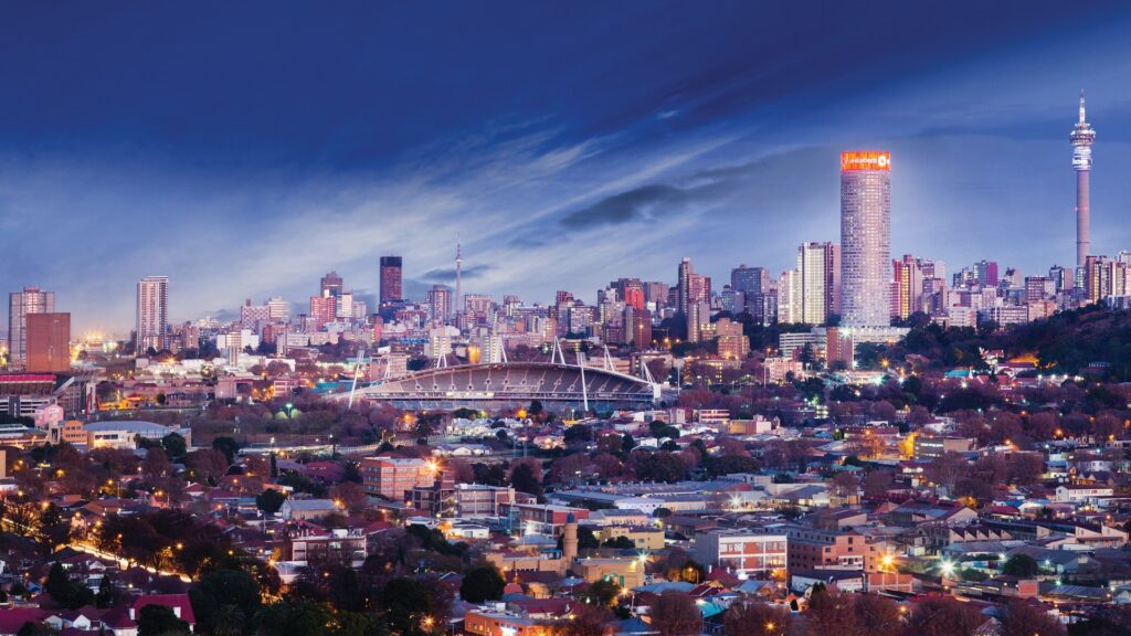 A panoramic view of Johannesburg skyline at sunset, featuring modern skyscrapers, urban buildings, and a vibrant orange sky.