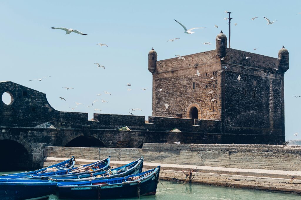 A historic coastal fortress or castle with traditional blue fishing boats moored in the foreground, and seagulls flying in the sky above. Essaouira